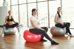 Positive pregnant ladies sitting on fit balls during prenatal training in spacious light gym at daytime and smiling while looking at camera