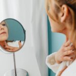 Hashimoto - przyczyny, objawy i sposoby leczenia choroby Woman in a bathrobe applying skincare while looking in the mirror.